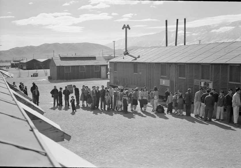 People of all ages wait in a line in front of a building at midday.