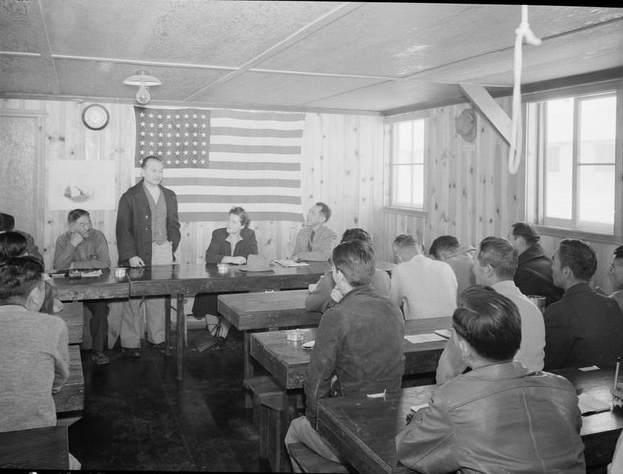 Roy Takeno, full-length portrait, standing behind a table, addressing a group with an American flag on the wall behind him.