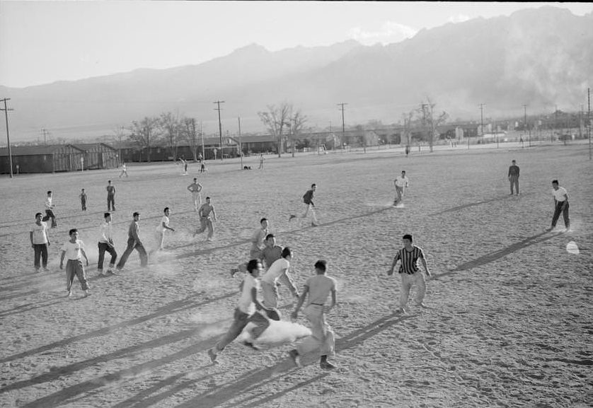 Players involved in a football game on a dusty field, buildings and mountains in the distance.