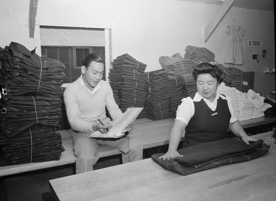 Bert K. Miura, seated on table with bundles of clothing, holding paper and pencil, with Toshiko Kadonada, folding clothing in front of him.