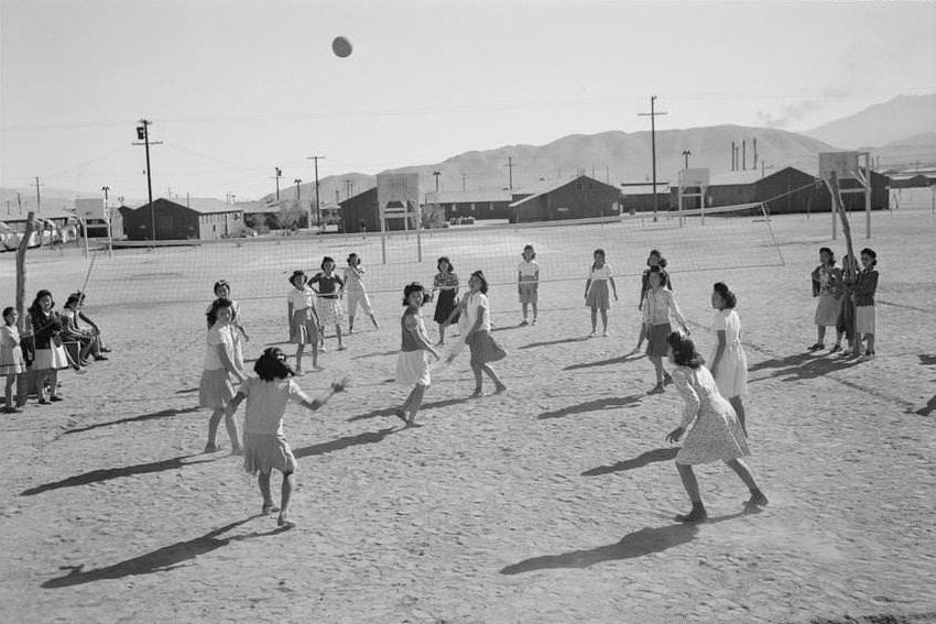 Japanese American women playing volleyball, one-story buildings and mountains in the background.