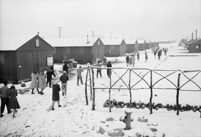 People walking through relocation center in snow past hand made wood fence.