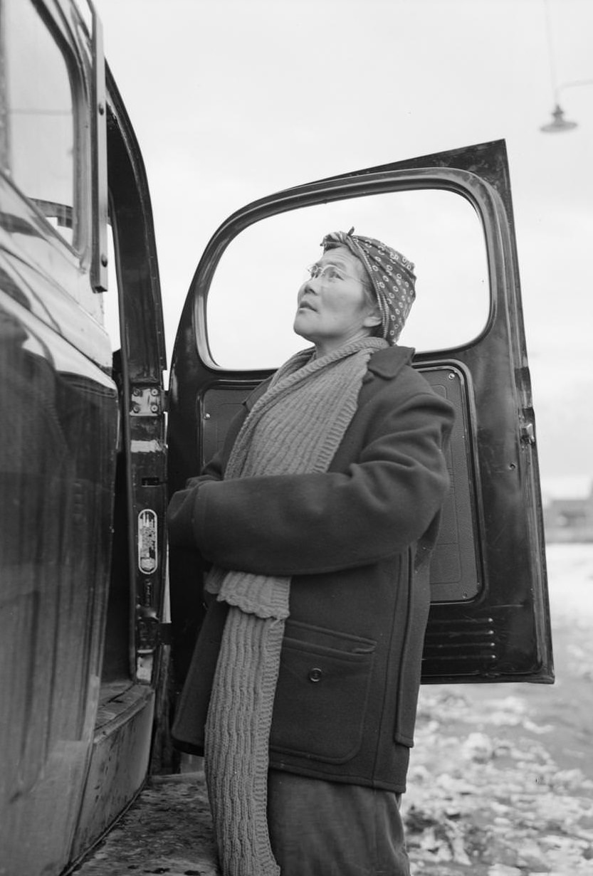 Mrs. Otsuno Nakaji, three-quarter length portrait, standing against the open door of an automobile, facing left.