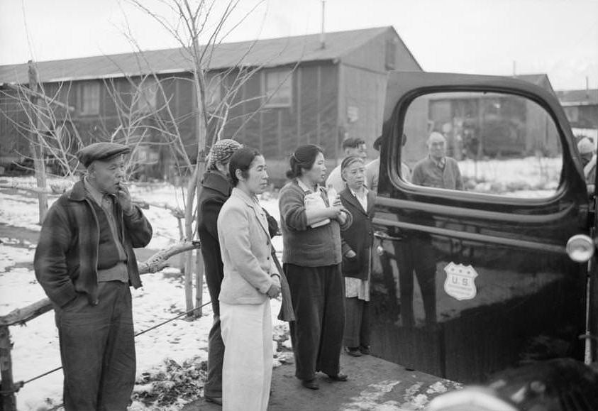 Group of people standing, facing right, near the open door of an automobile, buildings in the background.