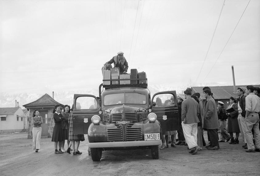 Man stands on top of bus loading luggage while a group of people gather to say farewell, guardhouse in the background.