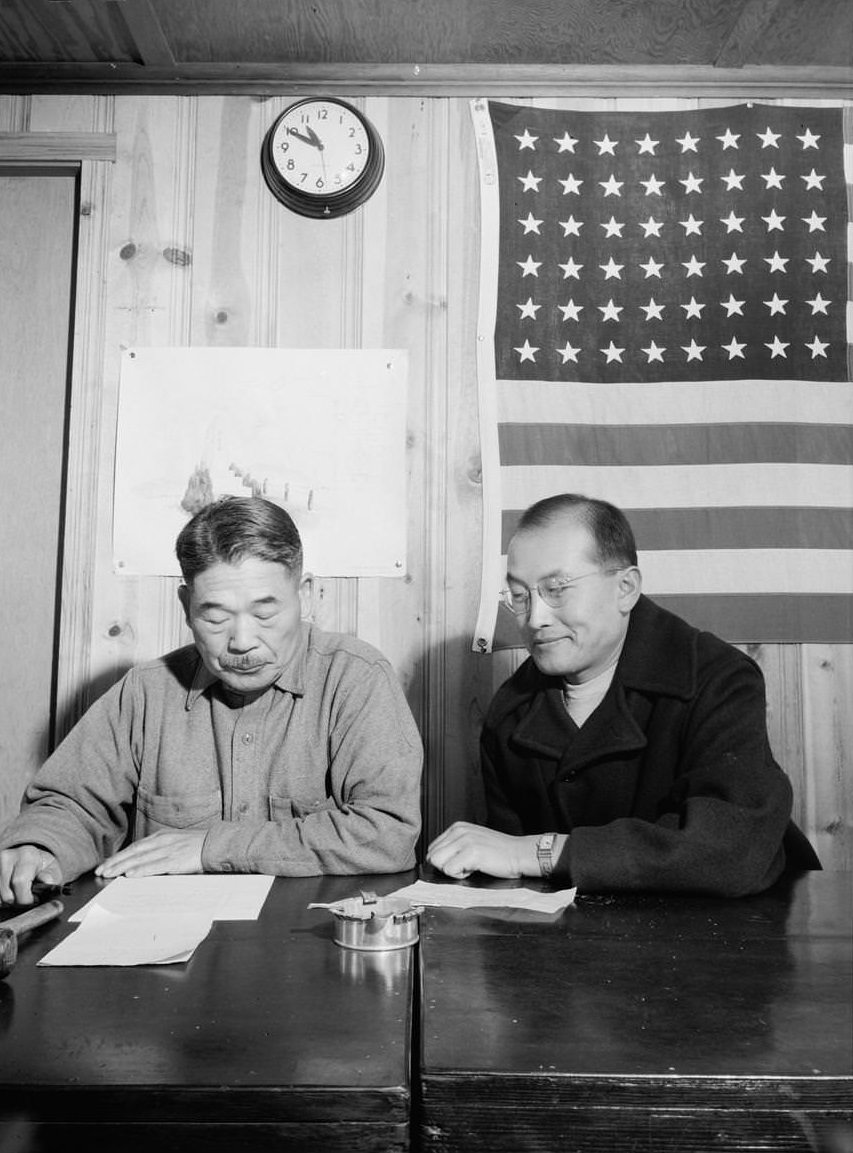 Roy Takeno (right), half-length portrait, seated at a table, next to the mayor who is reading papers, an American flag hangs on the wall behind them.