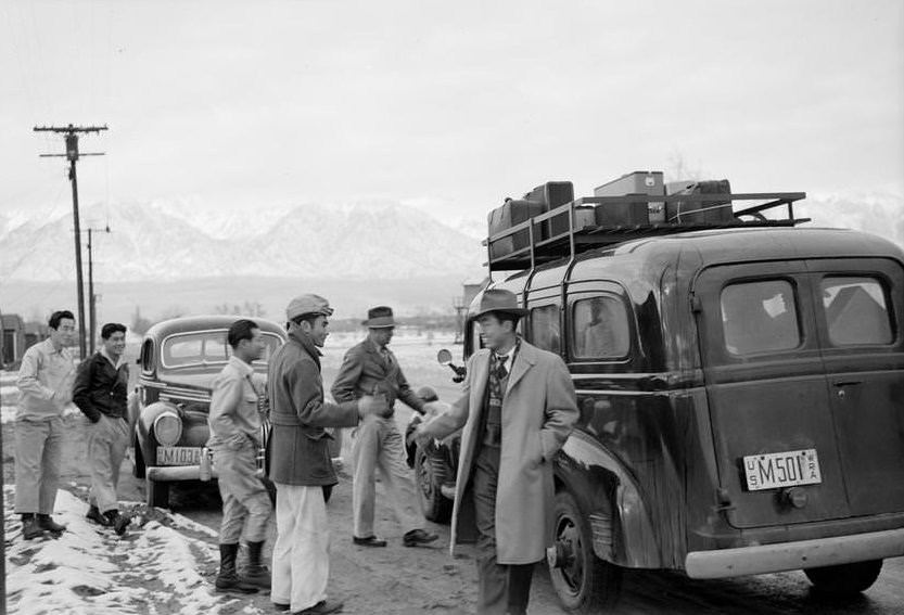 Group of men gathered around a bus packed with passengers and luggage say farewell, snow on the ground.
