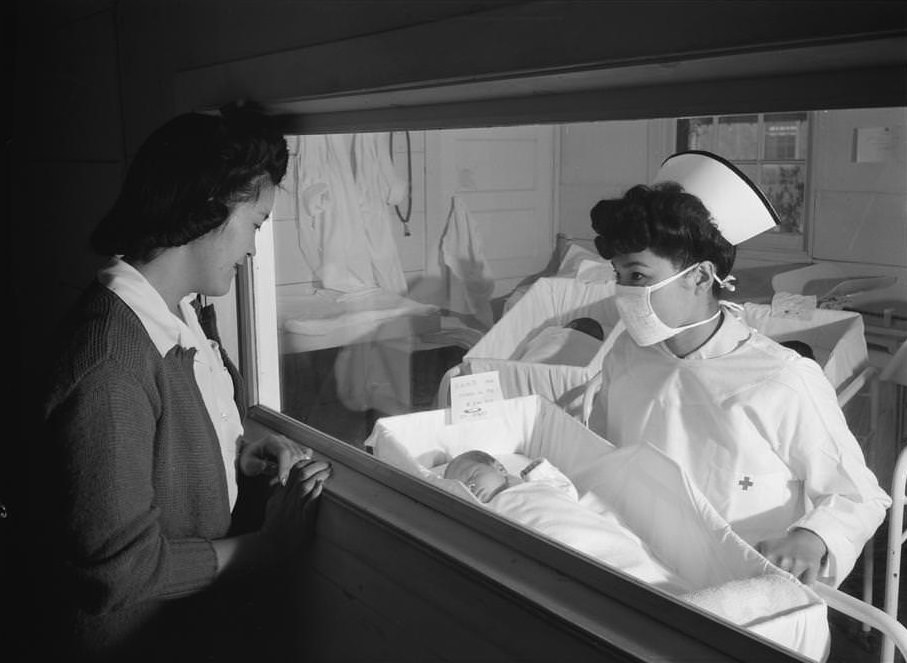 Woman looking through window at baby being held by nurse in maternity ward.