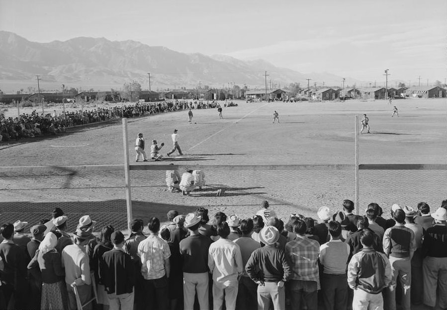 Japanese Americans observe an amateur baseball game in progress; one-story buildings and mountains in the background.