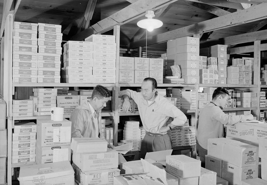 M. Ogi, S. Sugimoto, and Bunkichi Hayashi standing among shelves with boxes in warehouse.