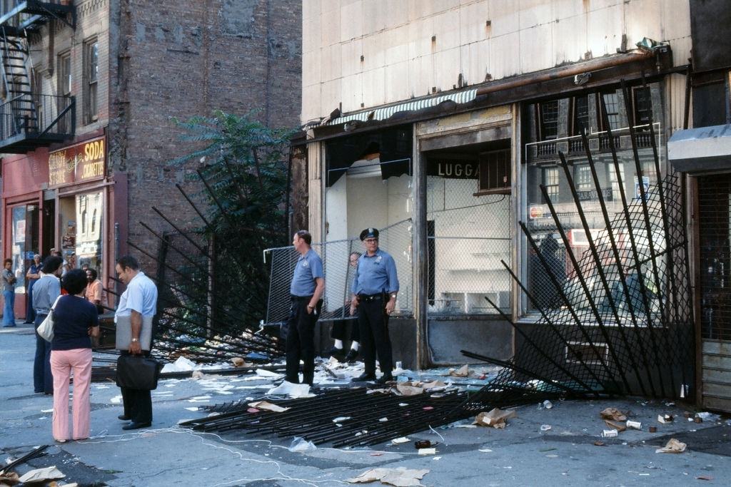 Police officers and passersby stand in front of a damaged store front, looted in the wake of the New York City blackout, New York City, 1977.