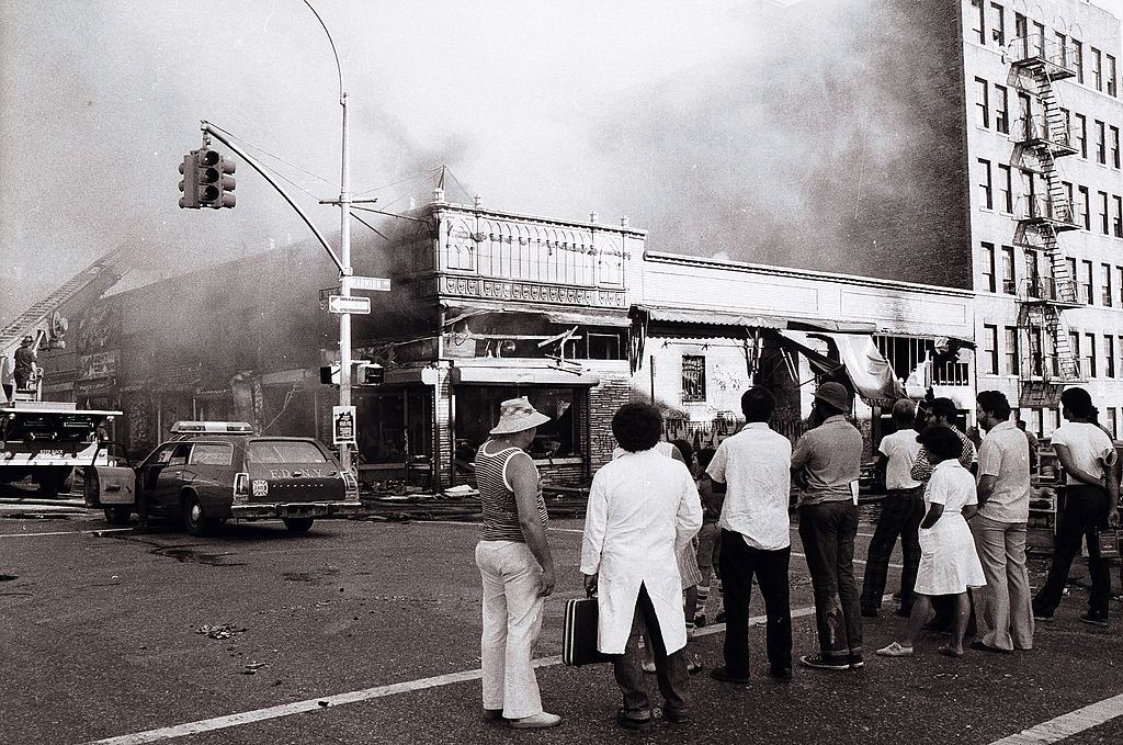 Az emberek tovább néznek, amint a tűzoltók véget vetnek az gyújtogatók kézműveinek a Marmion Ave-n, New York City-ben, 1977. július 13-án.