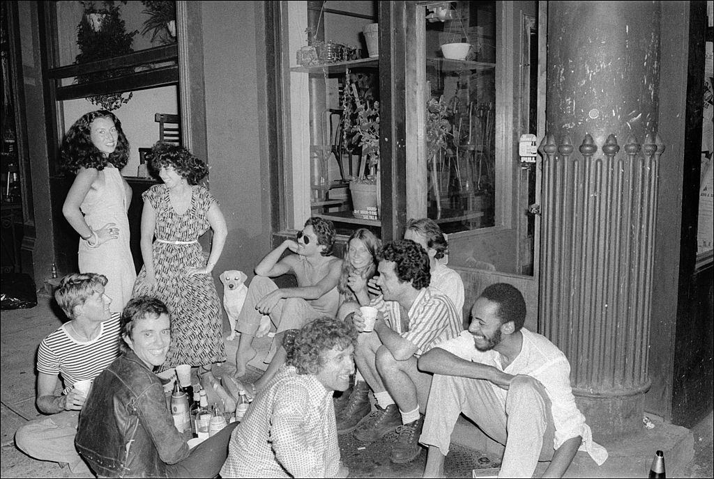 A group of people sits on the sidewalk and have a party outside the Food restaurant in SoHo, New York City, 1977.