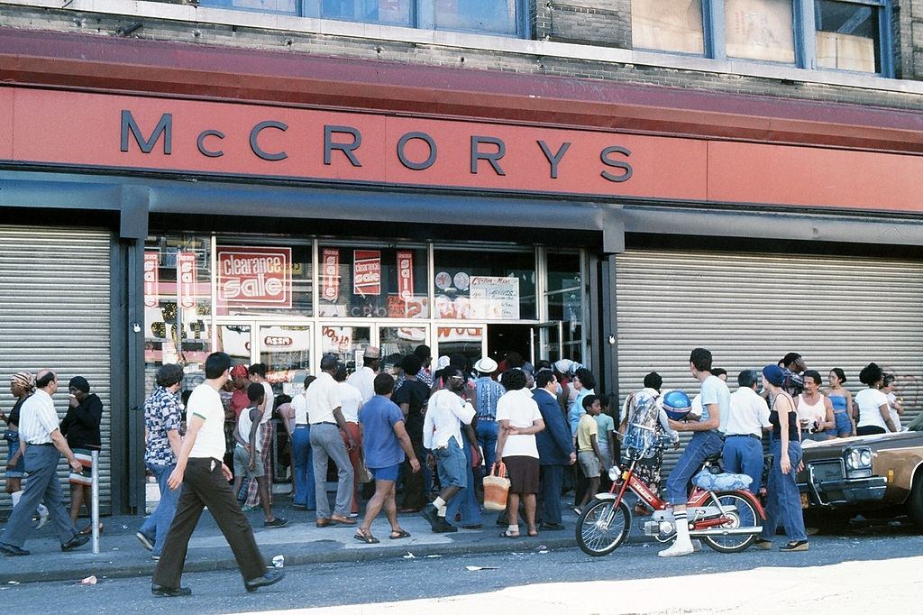 Police officers and passersby stand in front of a damaged store front, during the blackout, New York City, 1977.