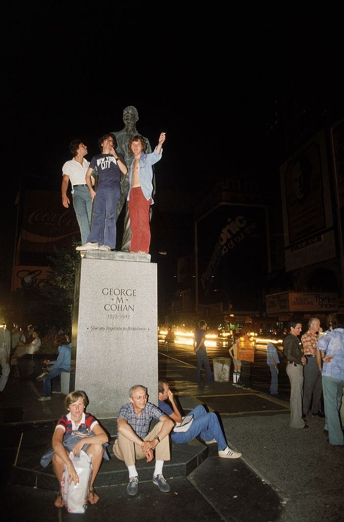 Az emberek összegyűlnek egy George M. Cohan zeneszerző emlékművéhez egy sötétebb Times Square-en egy áramkimaradás alatt, New York City, 1977. július 13.