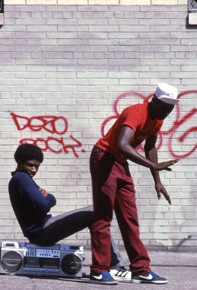 A young man breakdances on a city street while his friend sits on a boombox behind him, 1980s