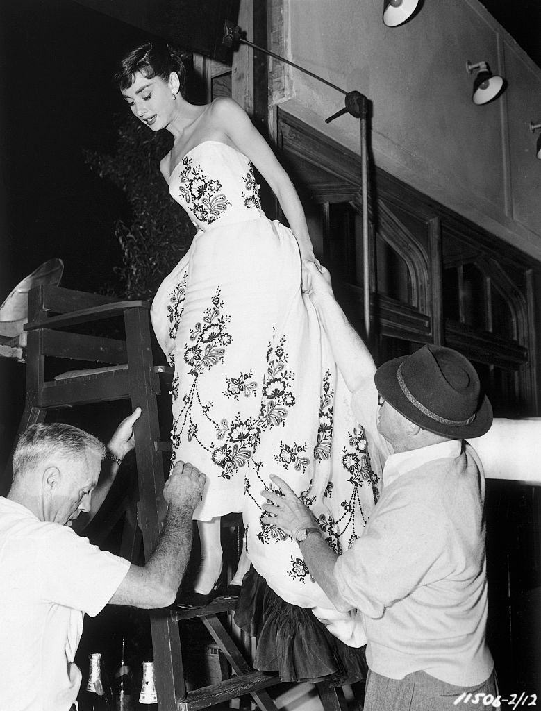 Audrey Hepburn standing halfway up a lifeguard's chair as she wears a flower-patterned dress.