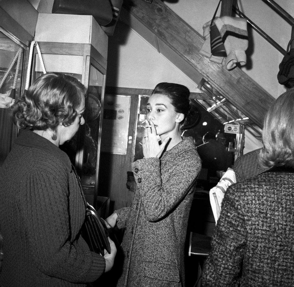Audrey Hepburn smoking a cigarette and talking to a woman in a leather shop. Rome, 1950s.