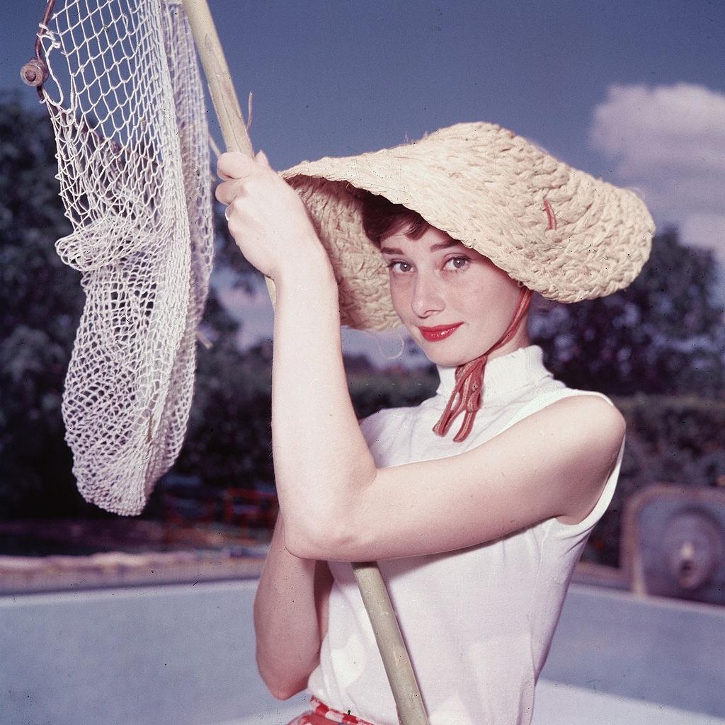 Audrey Hepburn in a peculiar hat and sleeveless blouse and holds a pool cleaning net beside a dry swimming pool, early 1950s.