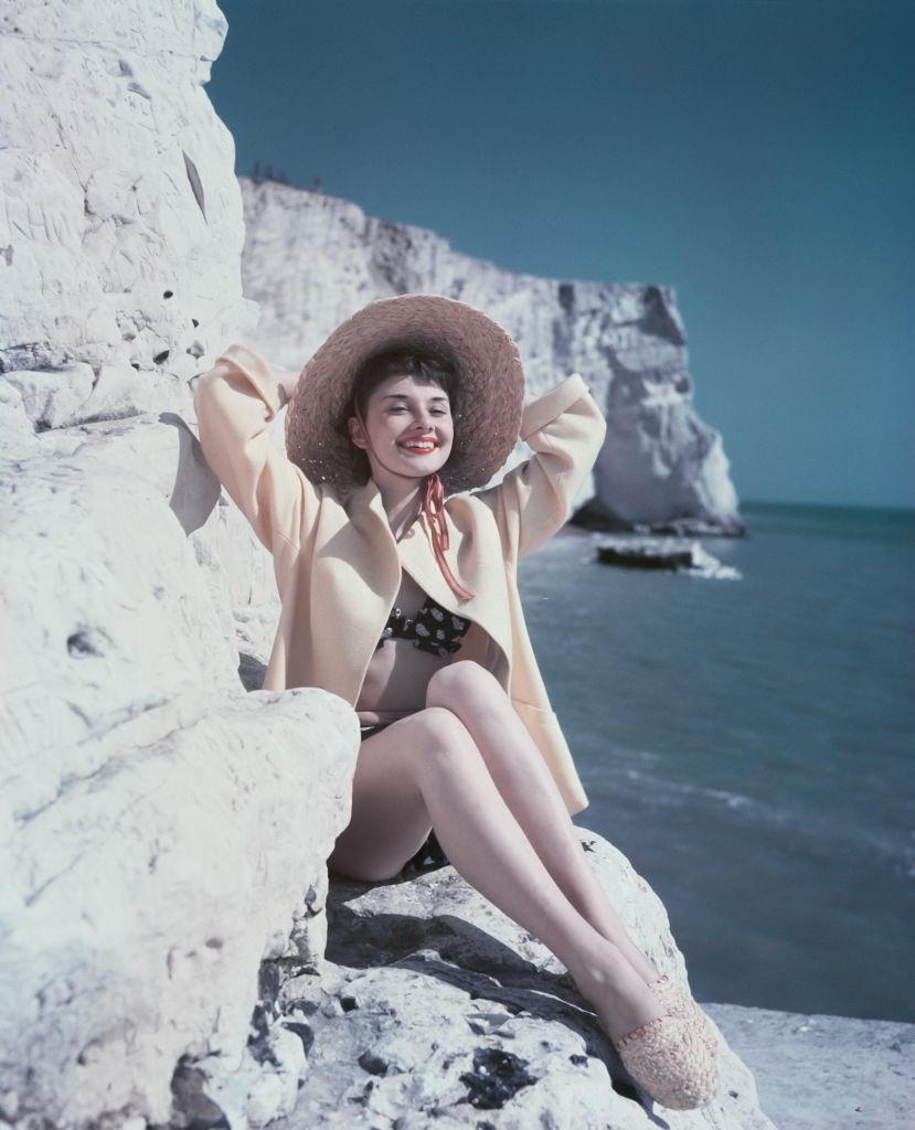 Audrey Hepburn relaxes by the coast as she sits on a rock wearing a hat and beachwear, 1951.