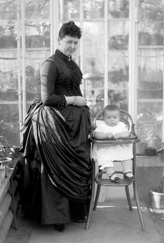 A well-to-do mother stands by her baby in a high chair, Belgium, 1901.