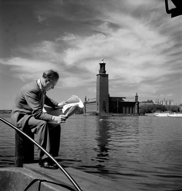A man sitting on a bollard and reading a newspaper. Stockholm City Hall and Riddarfjärden in the background.