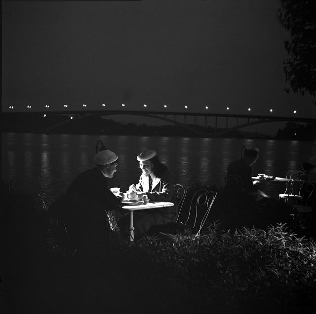 Two women sit at a table on an outdoor terrace in evening lighting, North Mälarstrand.