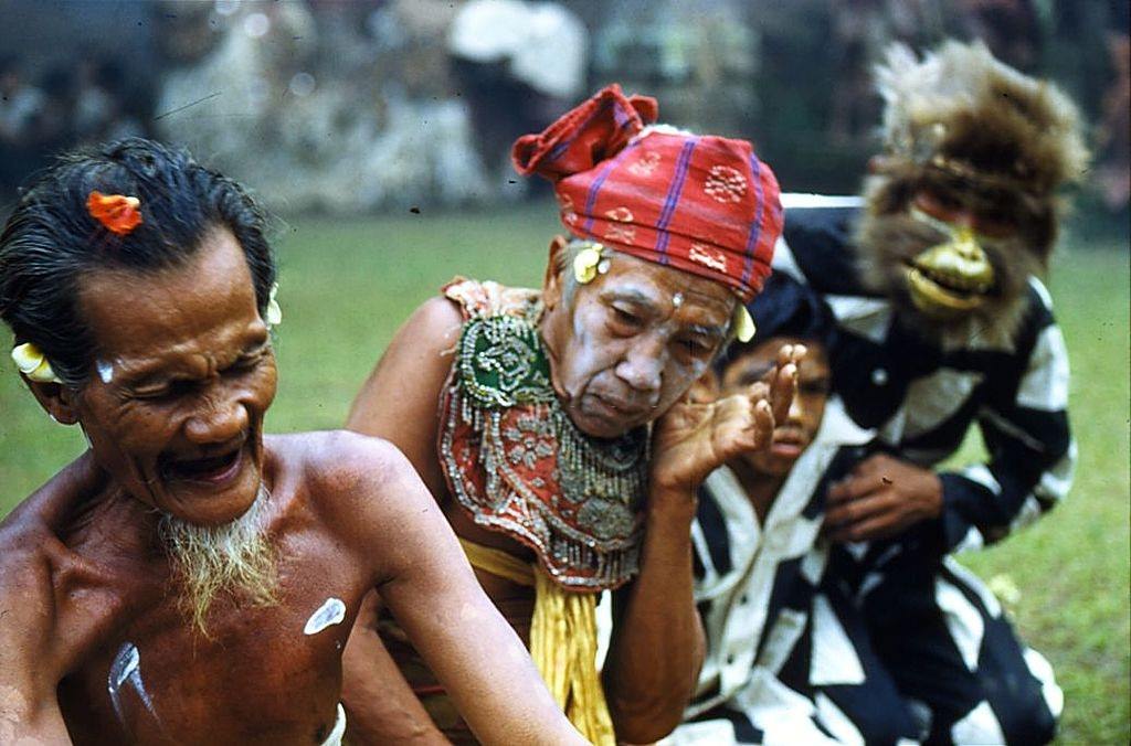 A devotee goes into a trance watched by an elderly man with a painted face and a man wearing a monkey mask, 1956.