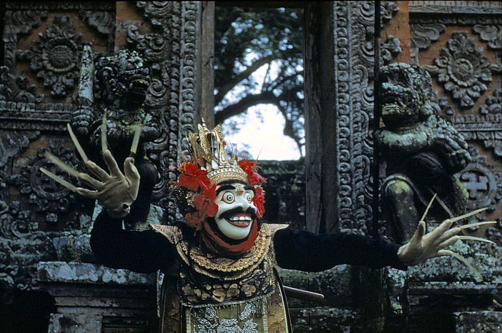 A Balinese 'topeng' or masked dancer in ceremonial headdress stands in front of ornately carved doors, 1956.