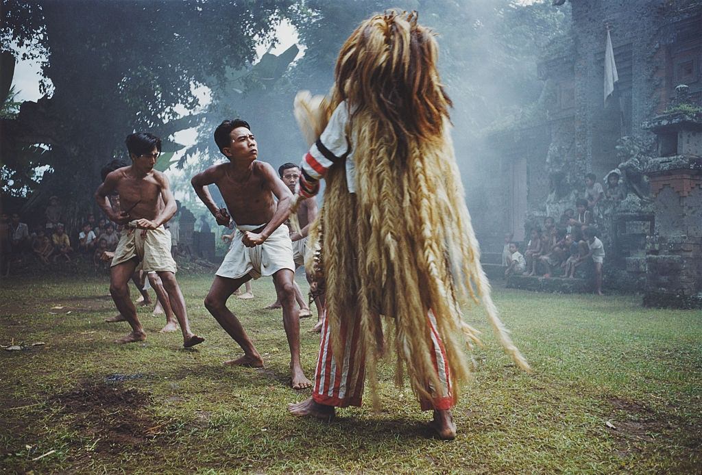 A human figure with hair reaching to its feet confronts a line of male dancers.The leonine figure is called the Barong. Bali, 1957