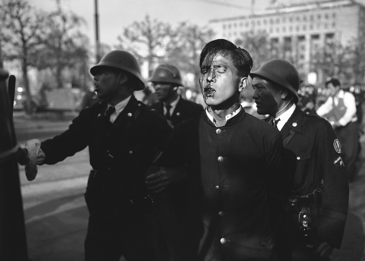 A dazed Japanese youth, his face bruised and bleeding, is led from the riot scene by a policemen after pro-communist demonstrators were dispersed near the imperial palace grounds in Tokyo on May 1, 1952.