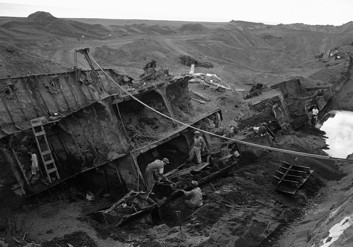 On the island of Iwo Jima, Japanese work crews cut up the wreckage of a naval vessel which was almost completely covered along the sandy beach on February 21, 1954.