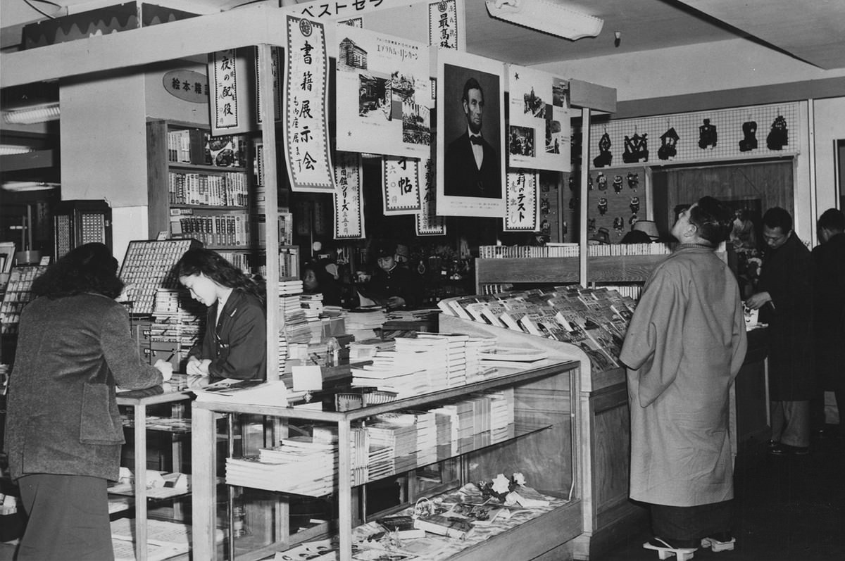 Interior of a Tokyo department store in 1959, where a Japanese man wearing Geta, traditional wooden footwear, looks up at a poster-sized portrait of Abraham Lincoln hanging with two other posters about Lincoln's life.