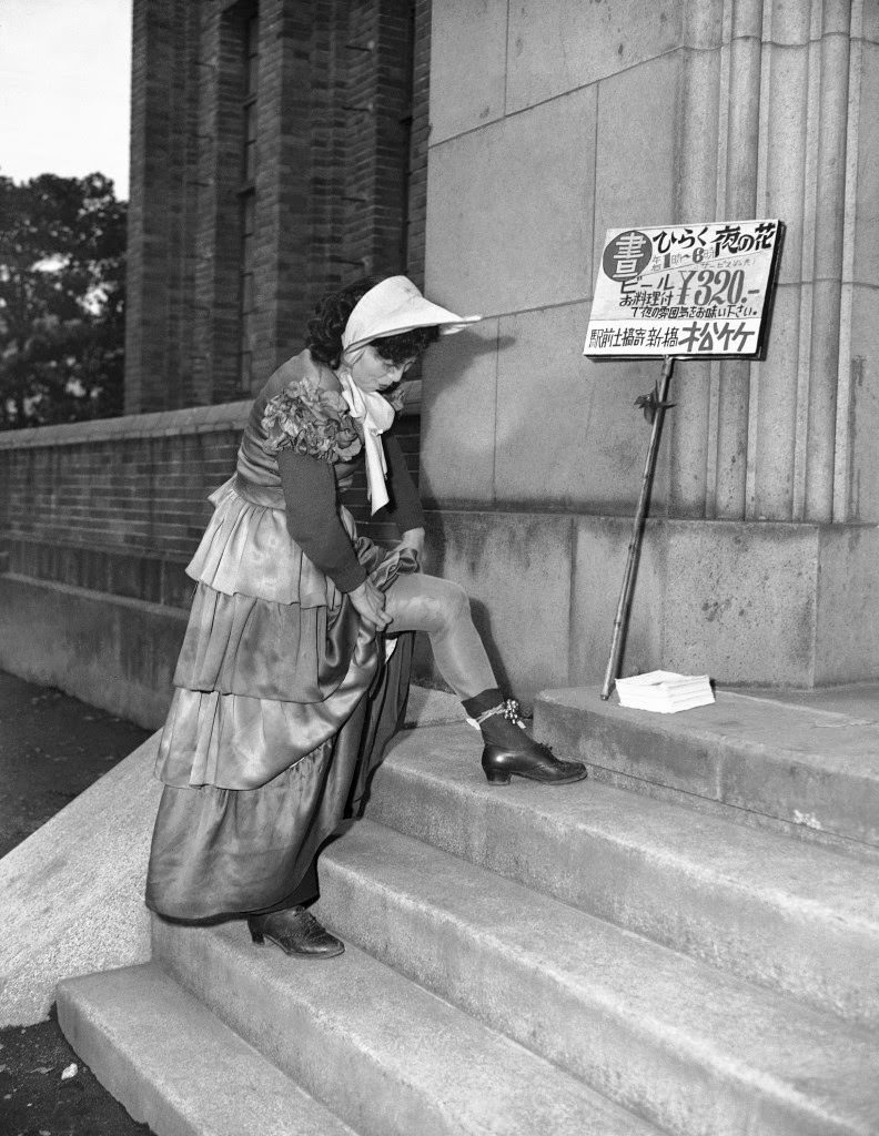 A sandwich girl carries a sign proclaiming a new type of club where folks who work nights can come and enjoy themselves during the day, stops to adjust the bells on her ankles in Tokyo, March 19, 1950.