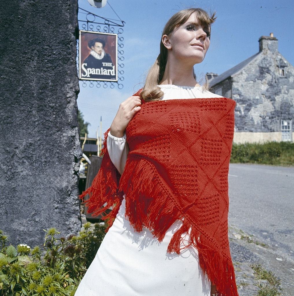 A young female model in a red knitted Spanish shawl over a white dress whilst standing outside The Spaniard Public House in Kinsale, Ireland in 1966.
