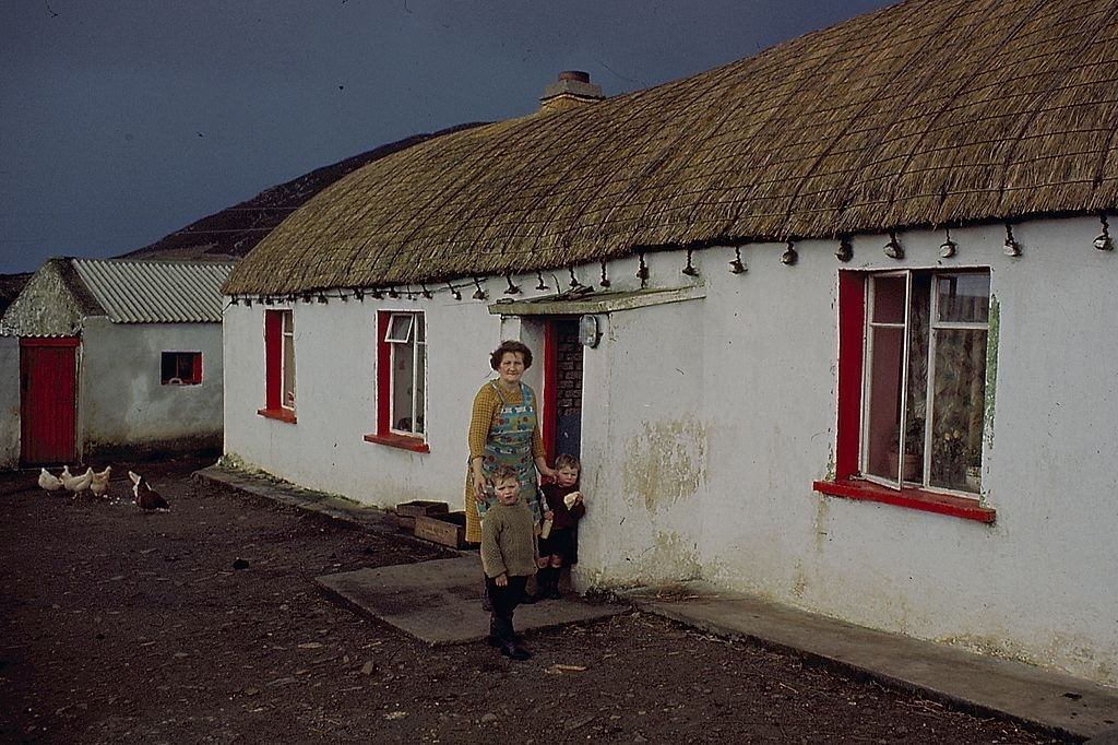 A woman stands with two small boys in front of a thatched cottage in County Donegal, Ireland in 1968.