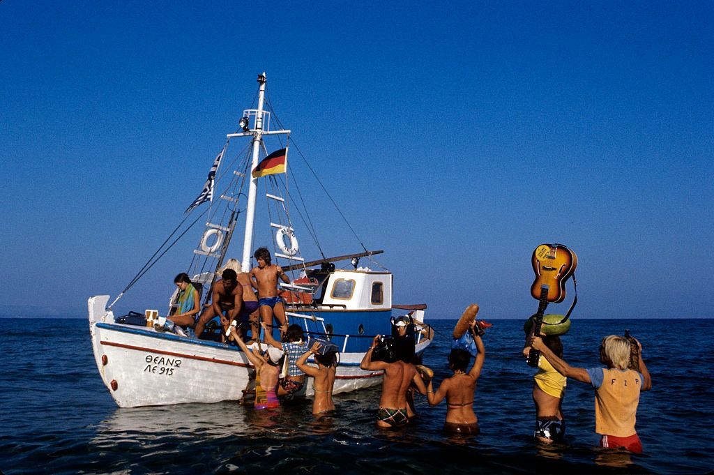 Tourists wade through the shallow water and board a fishing boat, Greece, 1974.