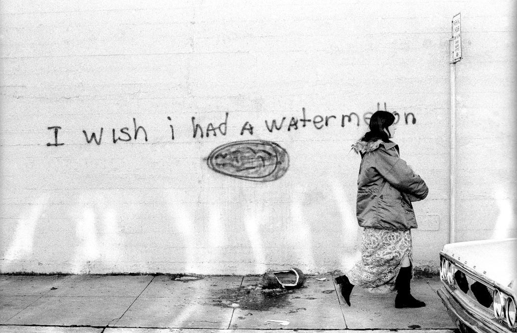 A woman walking by a message that says "I wish I had a Watermelon" with a crushed watermelon lays on the ground circa January, 1972.