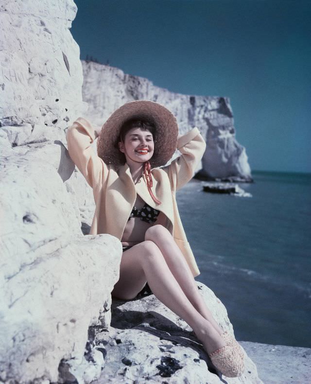 Audrey Hepburn sitting on a rock wearing a hat and beachwear, 1951.