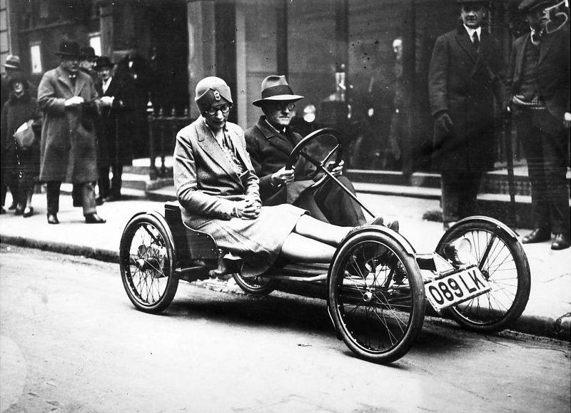 A man and a woman riding in an “Auto Red Bug”, an electric 2-seater runabout, in a London street.