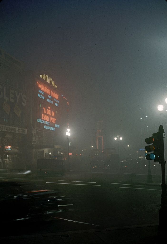 View along a foggy street where pedestrians pass a parked car, London, 1952.