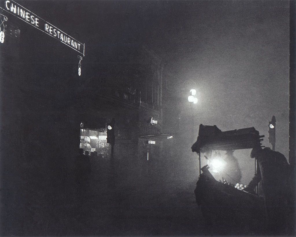 A foggy Piccadilly partially lit by the light from a fruit seller's stall, 1952.