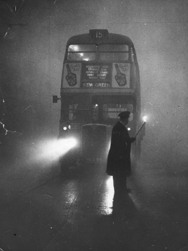 A man guiding a London bus through thick fog with a flaming torch.