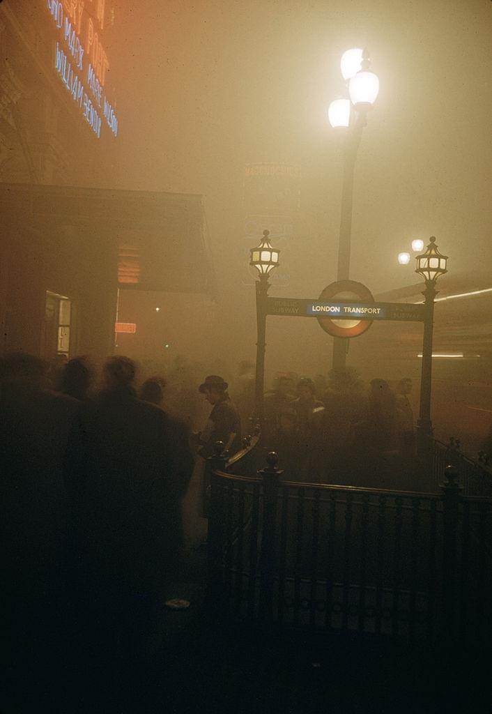 Pedestrian traffic on a foggy night outside a London Transport station and the London Pavillion cinema in Piccadilly Circus, London, 1952.