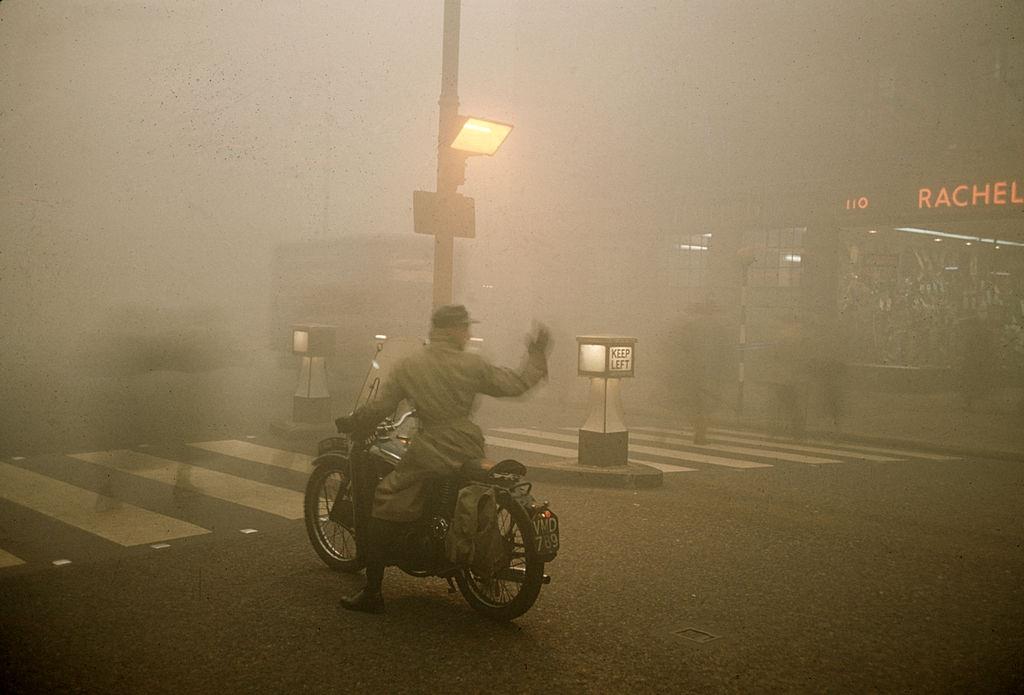 On a foggy night, a man on a motorcycle waits at a pedestrian cross-walk, London, 1952.