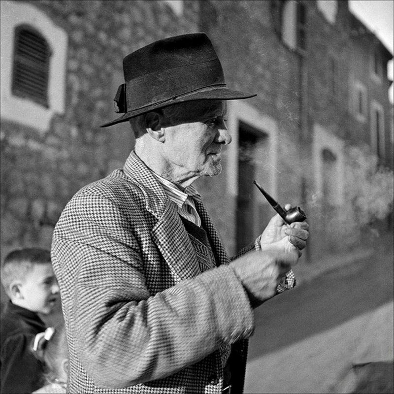 An old man smoking in the street and children in the background, 1956