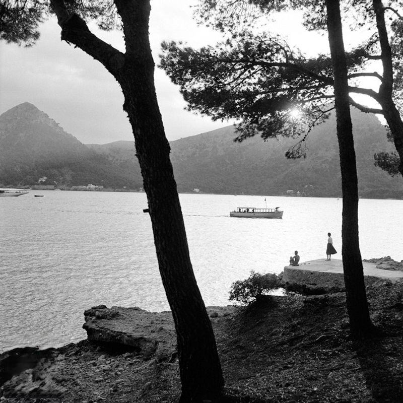 Two people by the sea with boat and mountains in the background, 1956