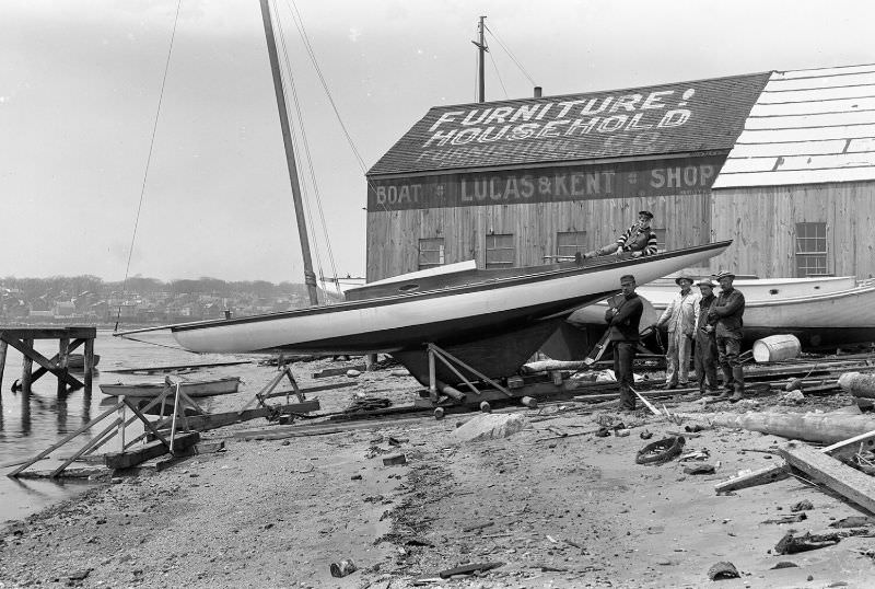 Men posed around sailboat. Lucas & Kent Boat Shop in the background, 1907