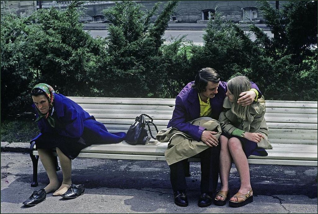 A couple share an intimate moment seated on a park bench whilst a woman next to them relaxes with her shoes off.