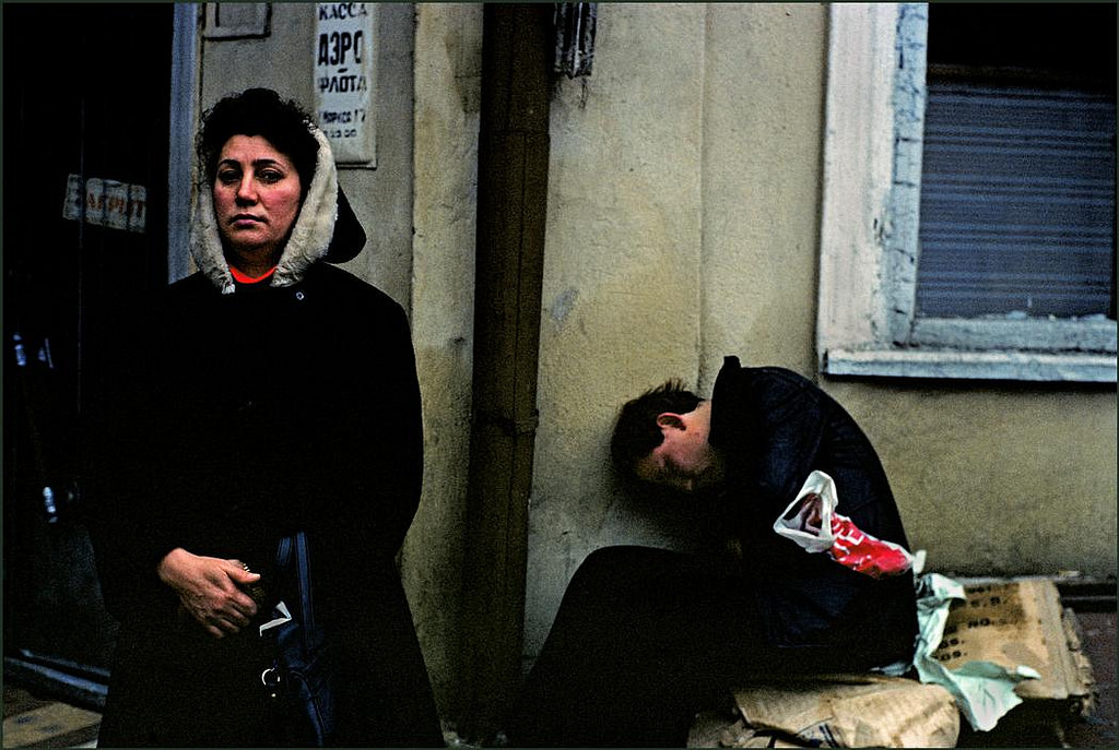 A woman waiting on the street ignores a drunk seemingly comatose behind her.